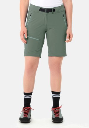 Person trägt olivgrüne knielange Shorts mit Seitentaschen, einen schwarzen Gürtel, schwarze Socken mit weißen Streifen und graue Wanderschuhe mit roten Schnürsenkeln.