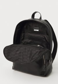 Zaino nero con finitura lucida, dotato di tasca frontale con zip e interno foderato con un motivo ripetuto "GUESS".