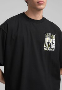 T-shirt nera in cotone a maniche corte, con una stampa grafica gialla e bianca sul lato sinistro del petto che recita "REPLAY MARTIN GARRIX."
