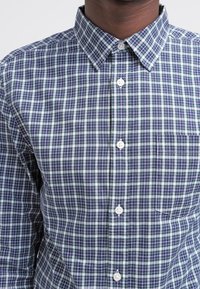 Skjorta med knäppning i rutigt mönster; har marinblå, grön och vit färg; inkluderar en enda bröstficka och en klassisk krage.