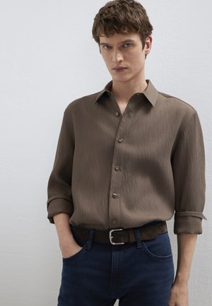 Junger Mann mit lockigem braunem Haar, der ein braunes, geripptes Hemd mit Knöpfen und aufgekrempelten Ärmeln sowie dunkelblaue Jeans mit braunem Gürtel trägt.