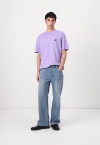 Purple cotton t-shirt with short sleeves and a small logo on the chest. Paired with light blue straight-leg jeans and black shoes.