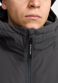 Chaqueta de pluma gris con collar alto, cremallera en contraste y tirador de marca. El material parece suave con un ligero brillo.
