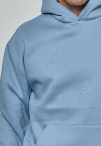 Homme portant un hoodie bleu clair avec une poche avant, visible des épaules au haut de la poitrine, montrant la barbe et la texture du tissu du hoodie.