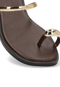 Sandal marrón con suela negra que presenta detalles metálicos dorados en el bucle del dedo y la correa del tobillo, vista en primer plano de la sección delantera.