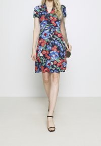 Donna che cammina indossando un abito floreale blu navy con fiori blu e rossi, tacchi a strappo neri e portando una piccola borsa a tracolla.