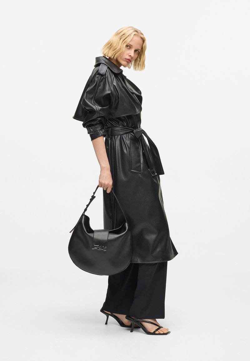 Vrouw in zwarte gestructureerde leren trenchcoat met een zwarte leren handtas, gekleed in zwarte wijde broek en zwarte sandalen met bandjes tegen een witte achtergrond.