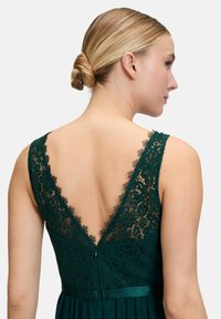 Emerald green dress featuring a deep V lace back, adjustable straps, a fitted bodice, and a flowing skirt with a satin waistband.