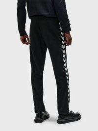 Ein Mann steht mit dem Rücken zu uns, trägt schwarze Hosen mit weißen Chevron-Streifen an den Seiten, schwarze Schuhe und ein schwarzes langärmeliges Oberteil.