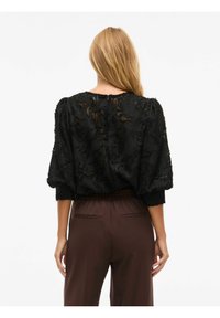 Svart teksturert bluse med puffede ermer, med et abstrakt mønster og rund halslinning. Kombinert med mørkebrune høy- midje bukser.