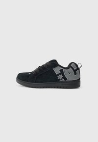 Laag uitgesneden zwarte suède sneaker met grijs DC-logo op de zijkant, zwarte veters en zwarte rubberen zool op een effen lichtgrijze achtergrond.