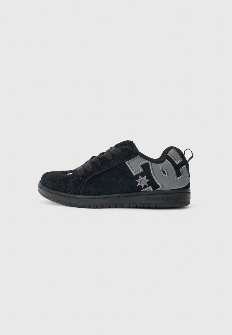 Laag uitgesneden zwarte suède sneaker met grijs DC-logo op de zijkant, zwarte veters en zwarte rubberen zool op een effen lichtgrijze achtergrond.