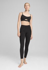 Crni atletski crop top s izrezom i tankim naramenicama, u kombinaciji s crnim hlačama visokog struka koje imaju glatku teksturu i šivane naglaske.