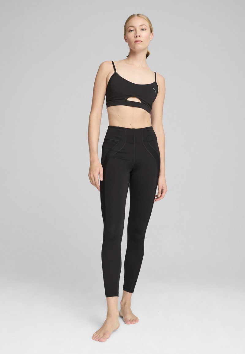 Crni atletski crop top s izrezom i tankim naramenicama, u kombinaciji s crnim hlačama visokog struka koje imaju glatku teksturu i šivane naglaske.