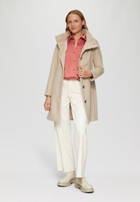 Manteau en laine beige avec un col côtelé, fermeture à boutons, superposé sur une chemise rose. Associé à un pantalon large blanc et des bottines beiges.