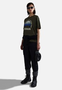 Camiseta verde oliva con gráfico de montaña, pantalones cargo negros, puños de calcetines grises de punto, botas negras con cordones y una gorra negra.