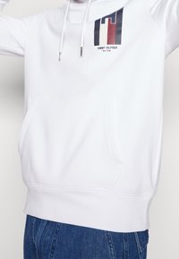 Felpa bianca con tasca a marsupio frontale, presenta un logo multicolore e testo sul petto. Tessuto morbido e liscio con cappuccio regolabile.