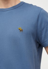 Μπλε μπλουζάκι με στρογγυλή λαιμόκοψη και μικρό κεντημένο καφέ ταράνδο με κίτρινα κέρατα στο αριστερό στήθος.