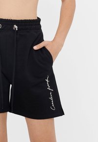 Černé bavlněné šortky s elastickým pasem, bočními kapsami a vyšitým textem bílou nití, který zní "Kreativní svoboda." Obsahují šňůrky na stažení v pase.