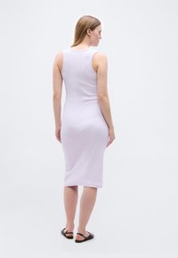 Γυναίκα που στέκεται με την πλάτη γυρισμένη, φορώντας αμάνικο μίντι φόρεμα σε ανοιχτό μοβ χρώμα και μαύρα σανδάλια με φλατ σόλα, σε απλό ανοιχτό φόντο.