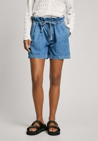 Shorts de mezclilla con cinturilla elástica y cordón. Color azul con un ajuste relajado. Llevados con sandalias negras con tachuelas doradas.
