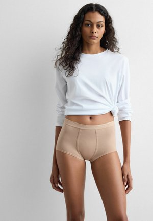 Mujer con cabello rizado, vistiendo una camisa blanca de manga larga anudada a la cintura y ropa interior beige de talle alto, de pie frente a un fondo liso.