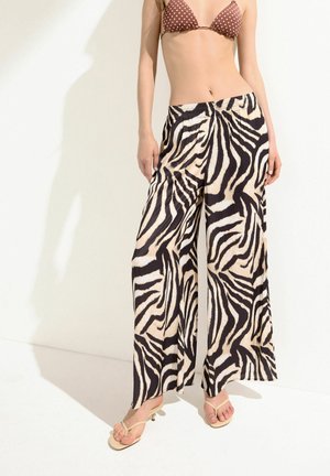 Femme portant un pantalon large imprimé zèbre beige et noir, un haut de bikini marron à pois et des sandales beiges à talons, debout sur fond blanc.