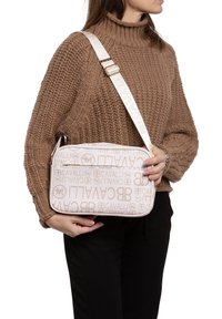 Borsa a tracolla beige con motivo del logo in rilievo, caratterizzata da una texture liscia e tracolla regolabile, tenuta da una persona che indossa un maglione lavorato a maglia.