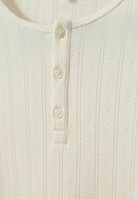 Krém színű ing texturált bordás mintával, lekerekített gallérral és elöl négy gombos légzőnyílással. Puha, könnyű anyag.