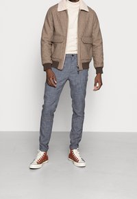 Hnědá texturovaná bomber bunda s krémovým límcem, šedé vzorované kalhoty a hnědé vysoké tenisky s bílou podrážkou.