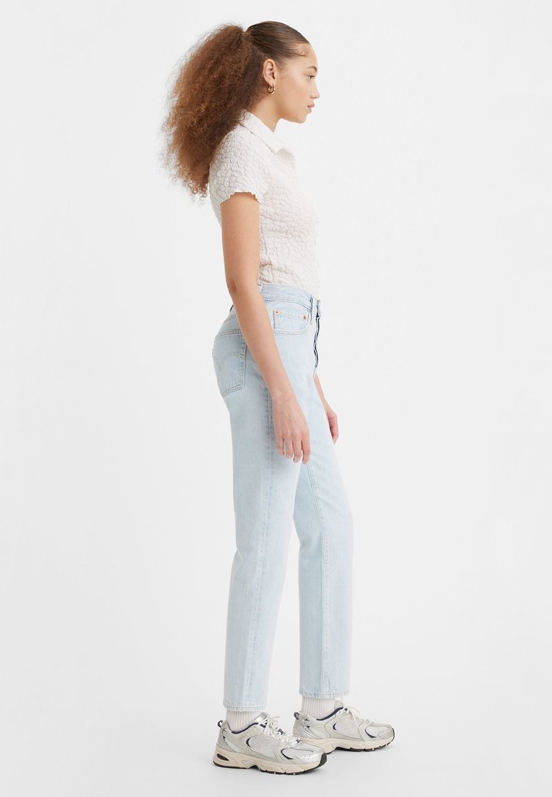 Jeans en denim bleu clair avec une coupe droite, associés à un haut blanc texturé à manches courtes et des baskets argentées à textures multiples.