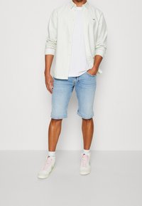 Ljusgrön skjorta över en vit t-shirt, tillsammans med ljusblå denimshorts och vita sneakers med rosa och gröna detaljer.