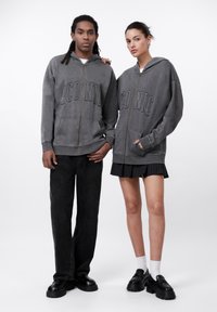 Siva zip-up dukserica s natpisom "IKONSKI", uparen s crnim hlačama širokih nogavica. Model s desne strane nosi plisiranu suknju i crne cipele s bijelim čarapama.