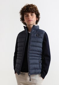 Marineblauer Puffer-Westen, gestepptes Design, hoher Kragen, Reißverschluss, Logo auf der Brust. Getragen über einem dunklen Pullover, kombiniert mit hellfarbenen Hosen.