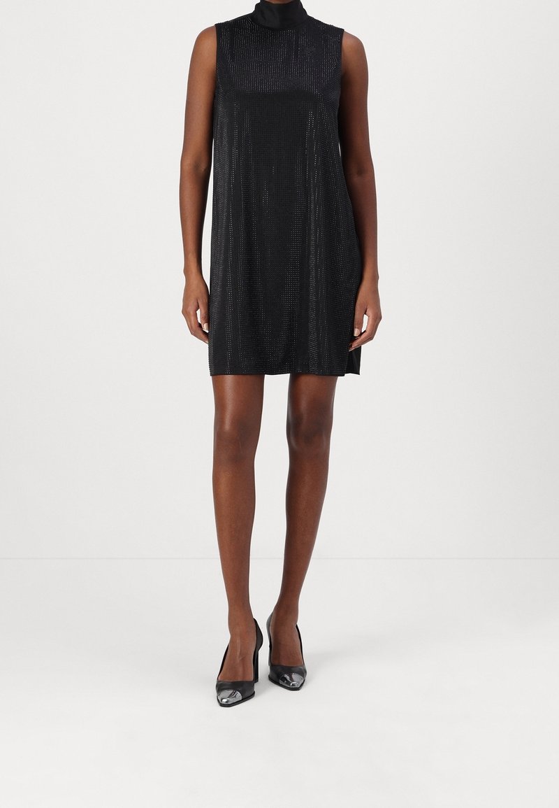 Femme portant une mini-robe noire texturée sans manches et des talons noirs à bouts pointus, debout devant un fond blanc uni.