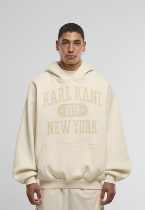 Fiatal férfi rövid hajjal, krémszínű, túlméretezett kapucnis pulóvert visel, amelyen a "KARL KANI 89 NEW YORK" felirat olvasható, egy sima háttér előtt áll.