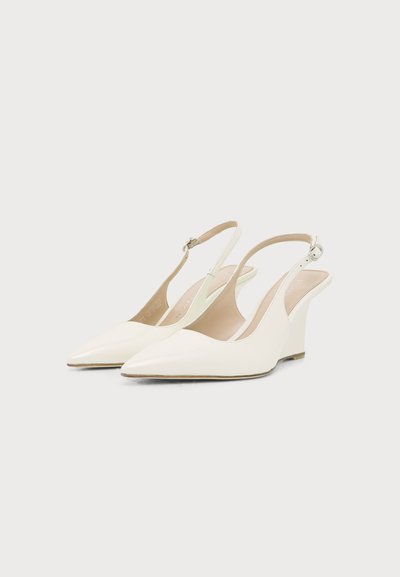 Paire d'escarpins compensés blancs à bout pointu, bride arrière et boucles argentées sur un fond clair uni.