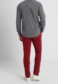 Mann trägt ein schwarz-weiß kariertes Hemd, rote Hose und weiße Turnschuhe, steht mit einer Hand in der Tasche und blickt weg.