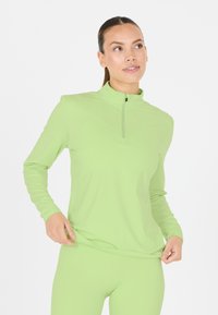 Femme aux cheveux attachés portant un haut zippé à manches longues vert citron et des leggings assortis, tenant l'ourlet de son haut.