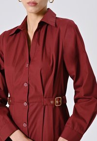 Donna che indossa un abito camicia bordeaux con cintura, bottoni dorati e fibbia, abbinato a orecchini a cerchio dorati.