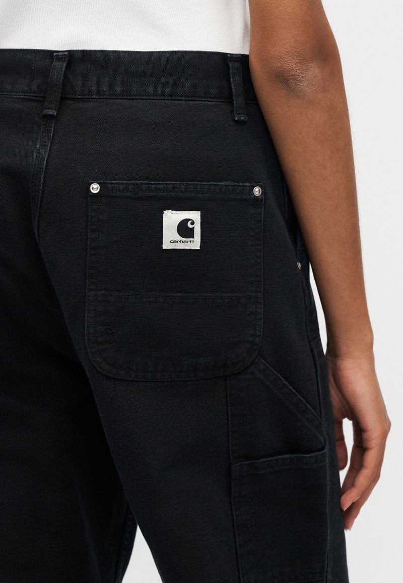 Pantalon cargo en coton noir avec une poche arrière ornée d'une étiquette blanche Carhartt. Des coutures visibles et des accessoires métalliques renforcent la durabilité.