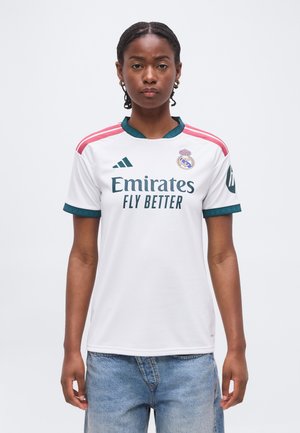 Kobieta z warkoczami, ubrana w białą koszulkę piłkarską Realu Madryt z logo „Emirates Fly Better” i niebieskie jeansy, stojąca na jednolitym tle.