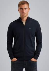 Mornarska zip-up dukserica od mekanog materijala, s rebrastim detaljima na ramenima i malim logom na prsima. Nosena je s bež majicom ispod.