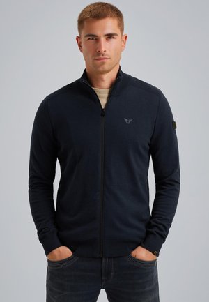 Navy zip-up sweater made of soft fabric, featuring ribbed shoulder accents and a small logo on the chest. Worn with a beige shirt underneath.