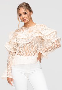 Femme aux cheveux blonds portant des boucles d'oreilles en perles, un chemisier en dentelle blanche avec des volants et un pantalon blanc sur un fond uni.