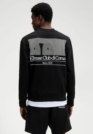 Mann trägt schwarzen Pullover mit weißem geometrischem Design und Text "Ellesse Club di Corsa Since 1959" auf der Rückseite, kombiniert mit schwarzen Shorts.