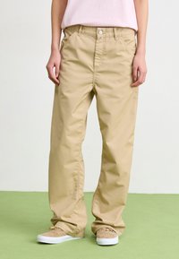 Beige vida byxor i bomull med knappstängning, med sidofickor och rak skärning, tillsammans med ljusbruna sneakers.