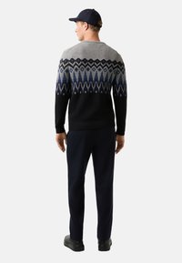 Mann mit schwarzer Mütze, schwarz-grau gemustertem Pullover, dunkelblauen Hosen und schwarzen Schuhen, der mit dem Rücken zu sehen ist, auf weißem Hintergrund.