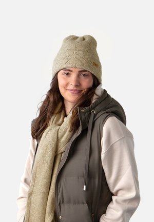 Bonnet en tricot beige avec un motif texturé, associé à une écharpe beige clair et un gilet sans manches matelassé vert olive, avec des accents argentés.