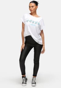 Hvid T-shirt med farverigt Smurfer-grafik, løs pasform, op-rullede ærmer. Sorte, slim-fit bukser lavet af et glat, skinnende materiale. Sorte sneakers.